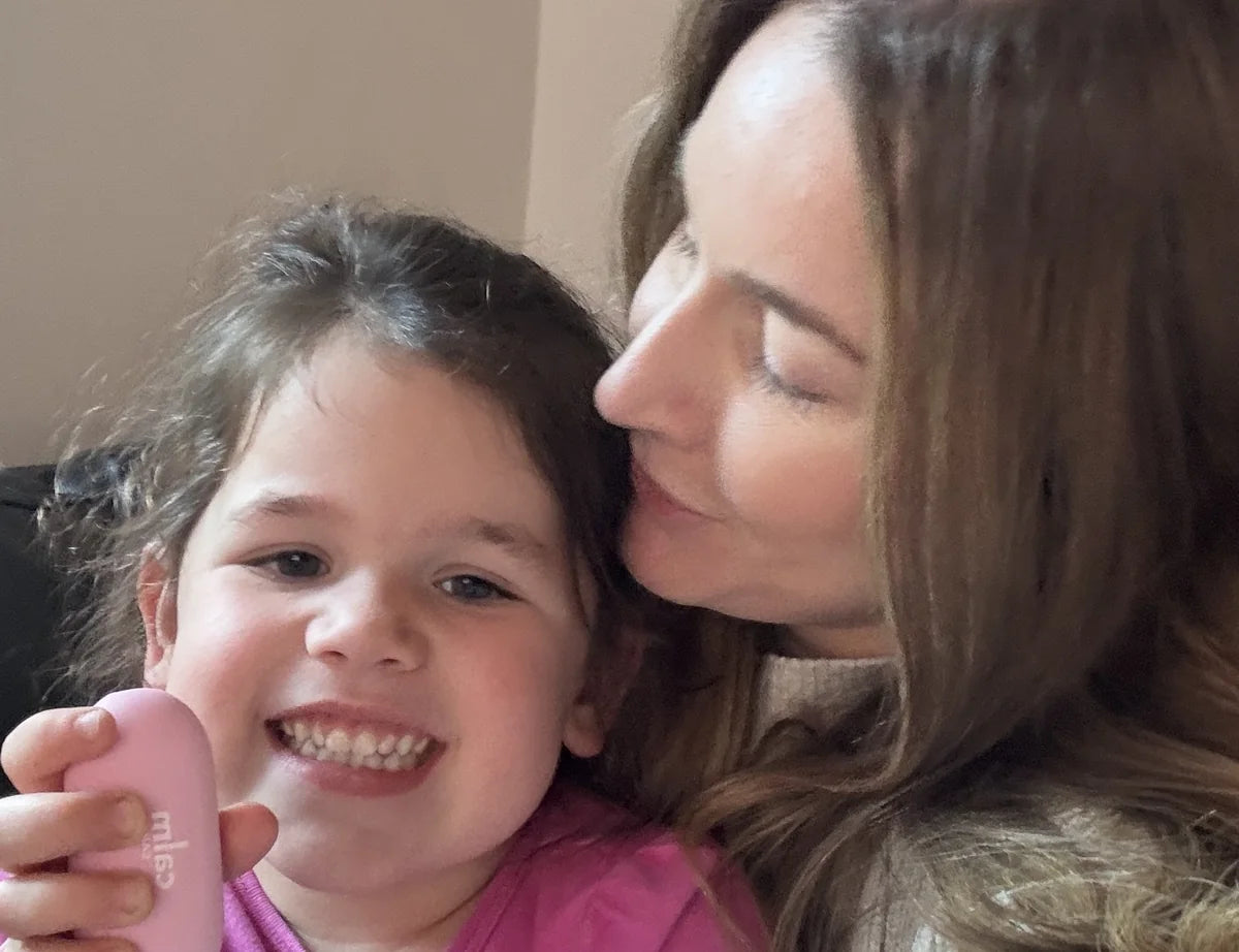 A mum kisses her smiling daughter, who is holding a pink CalmCarry